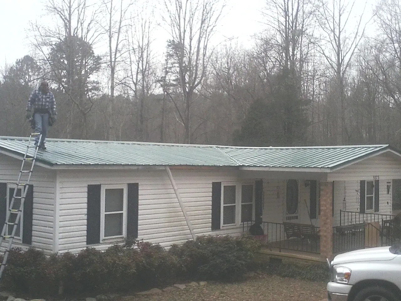 Worker on a ladder beside a new green metal roof on a ranch home in Haslett