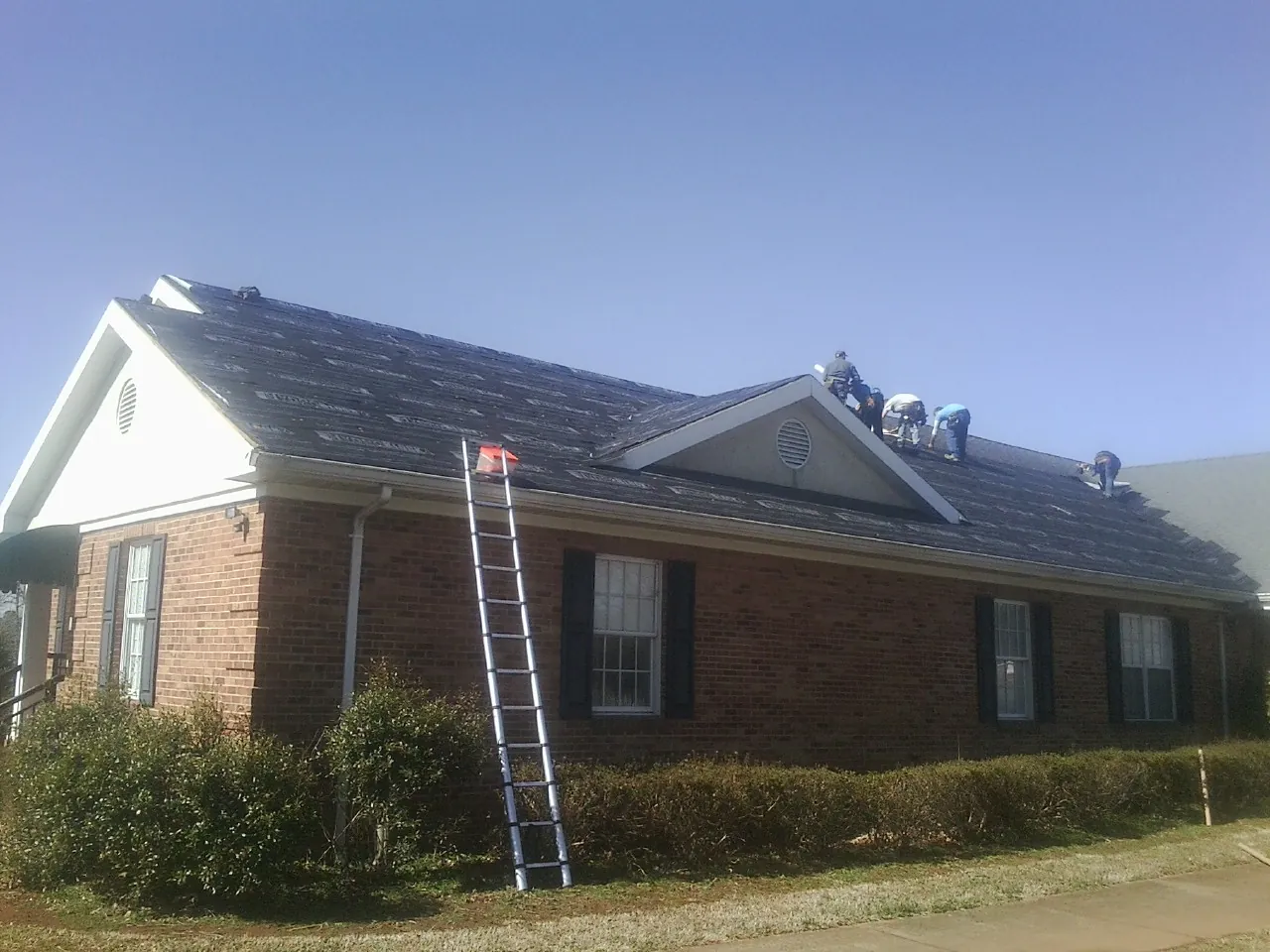 Crew tearing shingles off a brick ranch roof with a ladder set — Emergency Roof Repair work by our crew in Haslett