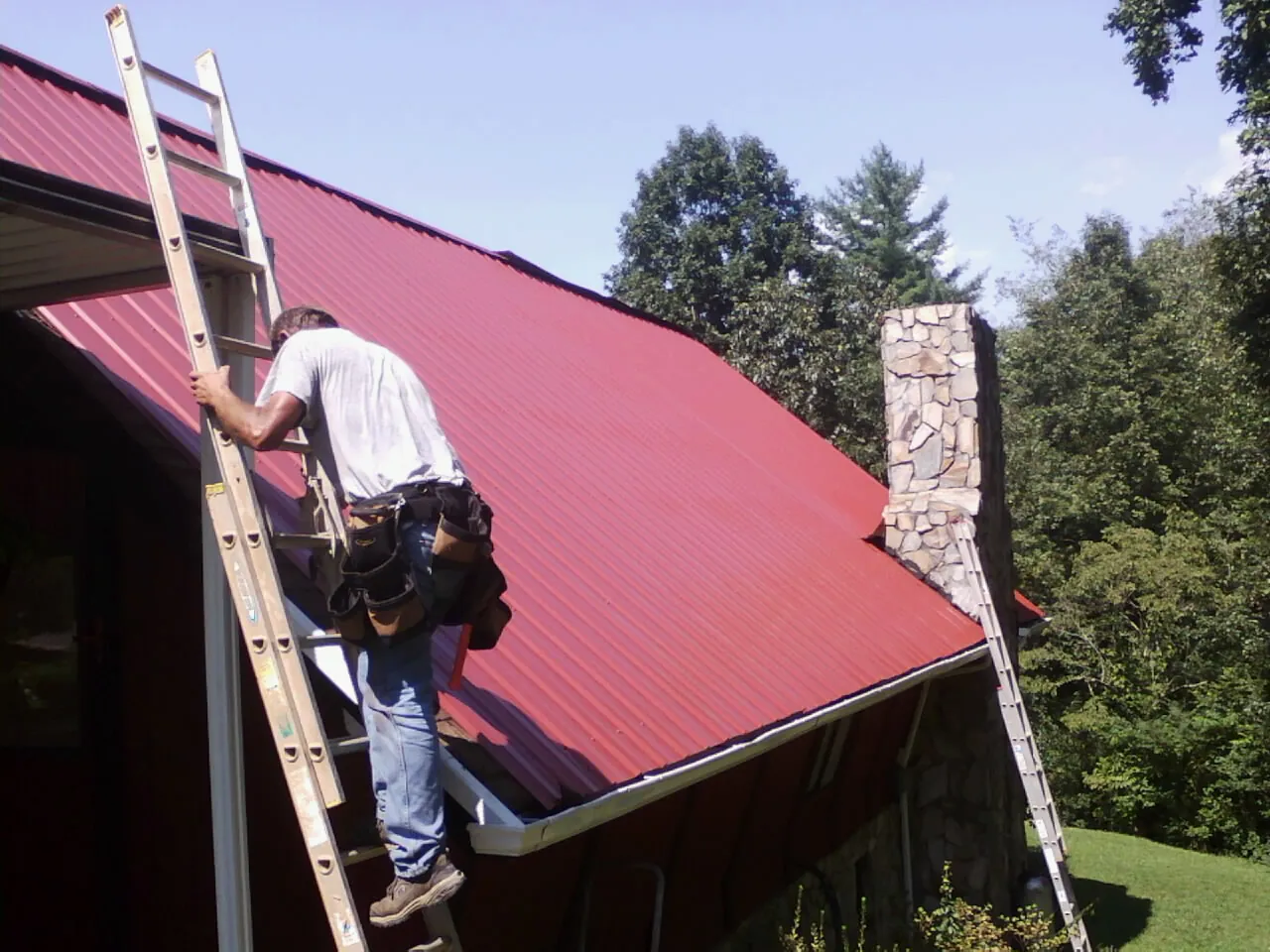 Roofer on a ladder beside a red metal roof and stone chimney — Soffit & Fascia Repair by Roofing Brothers in Haslett